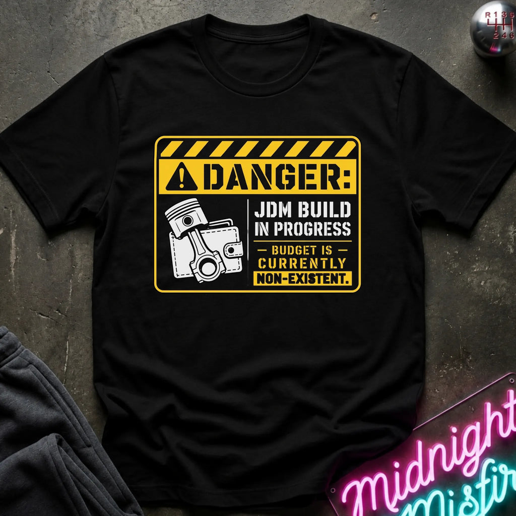 A black short-sleeve graphic T-shirt laid flat on a dark textured surface. The shirt features a yellow and white warning-sign style print with the text 'DANGER: JDM BUILD IN PROGRESS - BUDGET IS - CURRENTLY NON-EXISTENT'.