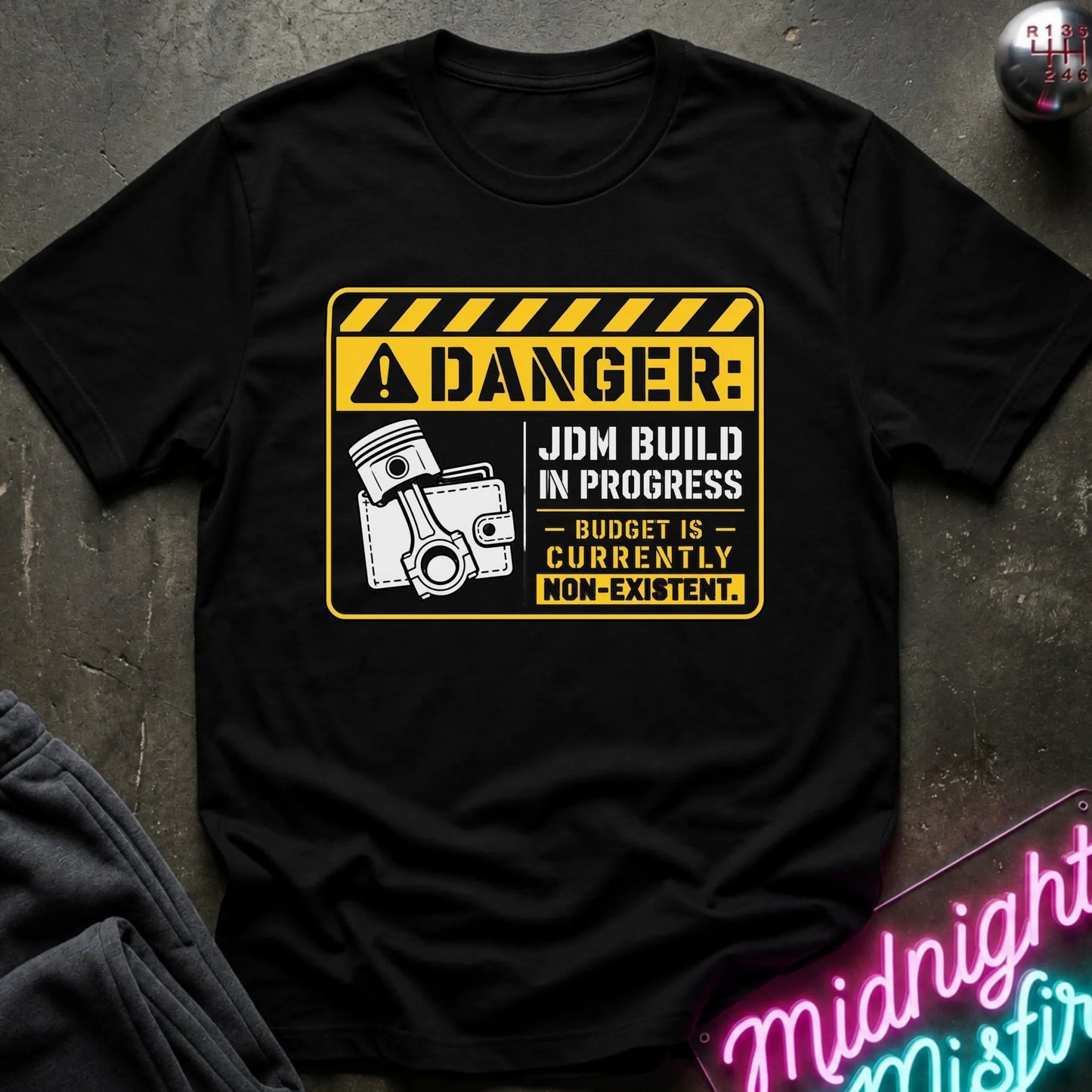 A black short-sleeve graphic T-shirt laid flat on a dark textured surface. The shirt features a yellow and white warning-sign style print with the text 'DANGER: JDM BUILD IN PROGRESS - BUDGET IS - CURRENTLY NON-EXISTENT'.