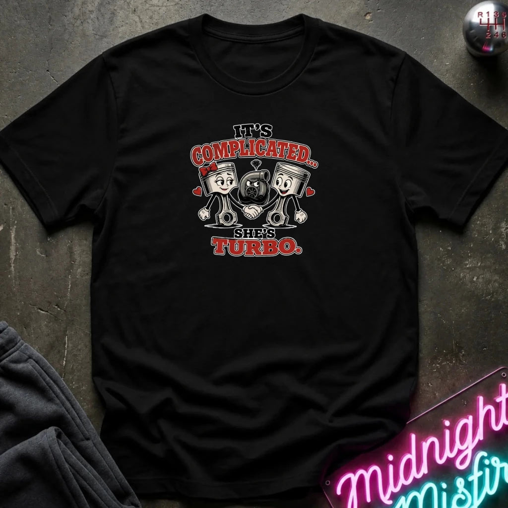 A black short-sleeve graphic t-shirt laid flat on a dark textured surface. The graphic features two cartoon pistons with hearts and the text 'IT'S COMPLICATED.