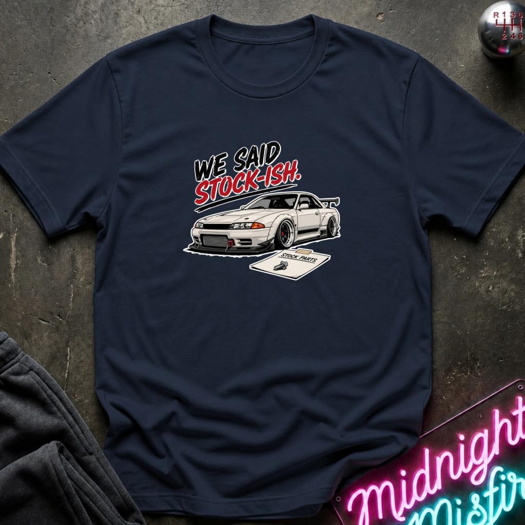 A navy blue graphic t-shirt laid flat on a dark textured surface. The shirt features a white sports car and the text 'WE SAID STOCK-ISH!' in stylized black and red lettering.