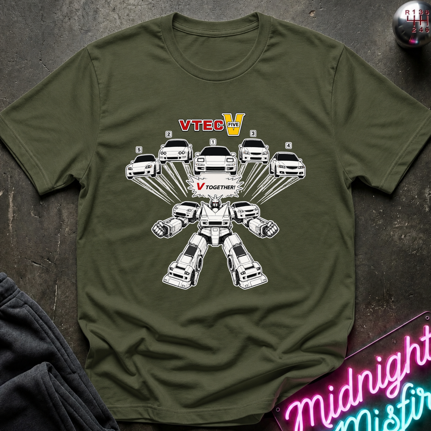 An olive green short-sleeve t-shirt with a graphic print of white cars transforming into a robot, with the text 'VTECH' and 'V TOGETHER'. The shirt is laid flat on a dark textured surface with a neon sign and dark clothing partially visible.