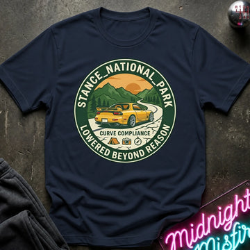 A navy blue graphic t-shirt laid flat on a dark textured surface. The shirt features a circular green and yellow graphic in the center depicting a yellow sports car on a winding road with mountains and an orange sunset, surrounded by the text 'STANCE NATIONAL PARK' and 'LOWERED BEYOND REASON'.
