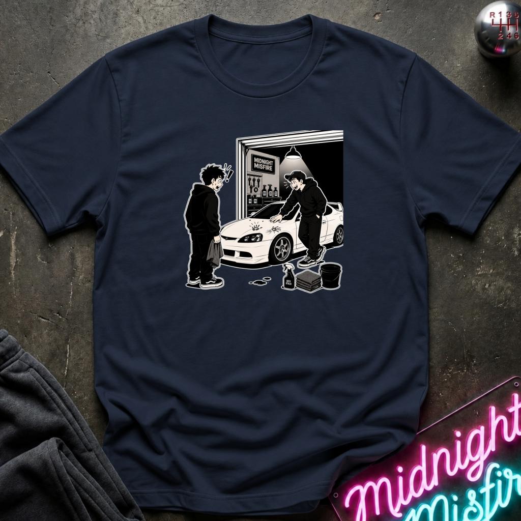 A navy blue short-sleeve t-shirt laid flat on a dark textured surface, featuring a black and white graphic print of two characters standing by a white sports car in front of a garage door.