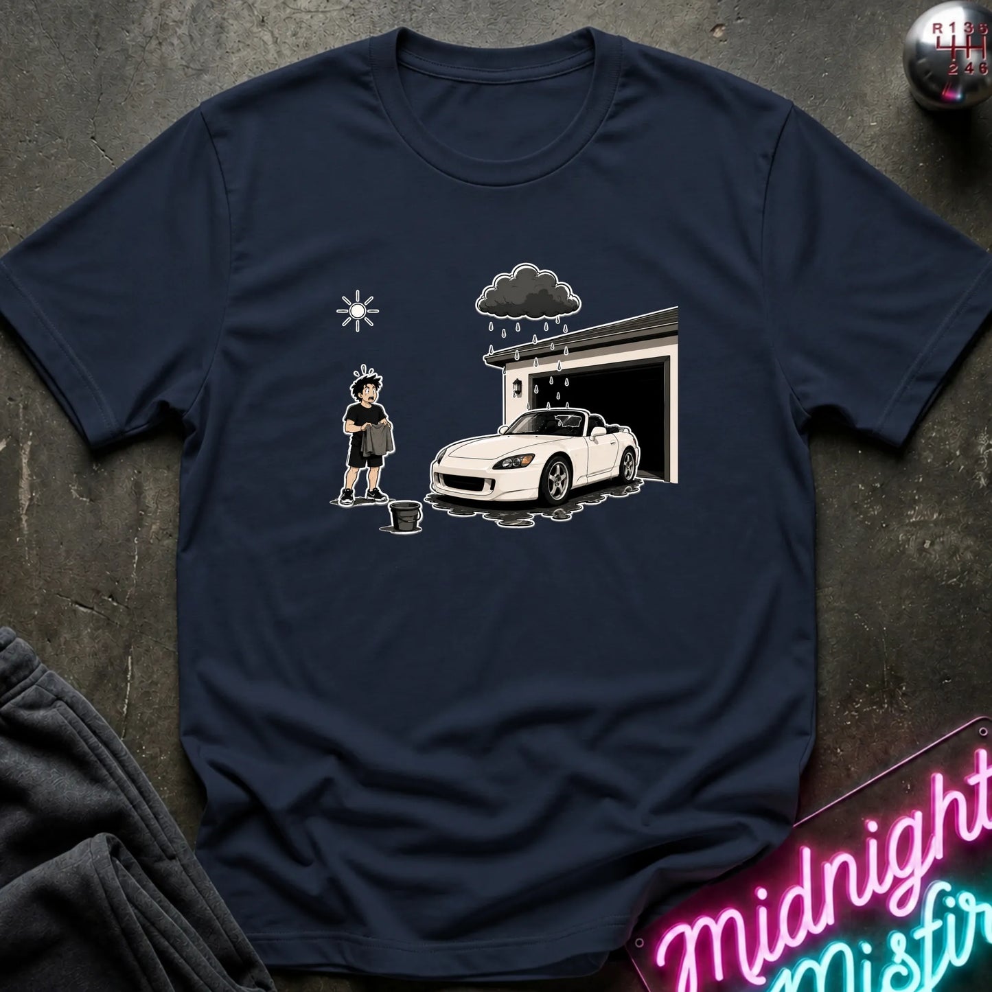 A navy blue short-sleeve t-shirt laid flat on a dark textured surface, featuring a cartoon graphic of a person washing a white convertible car in front of a garage under a dark raincloud.