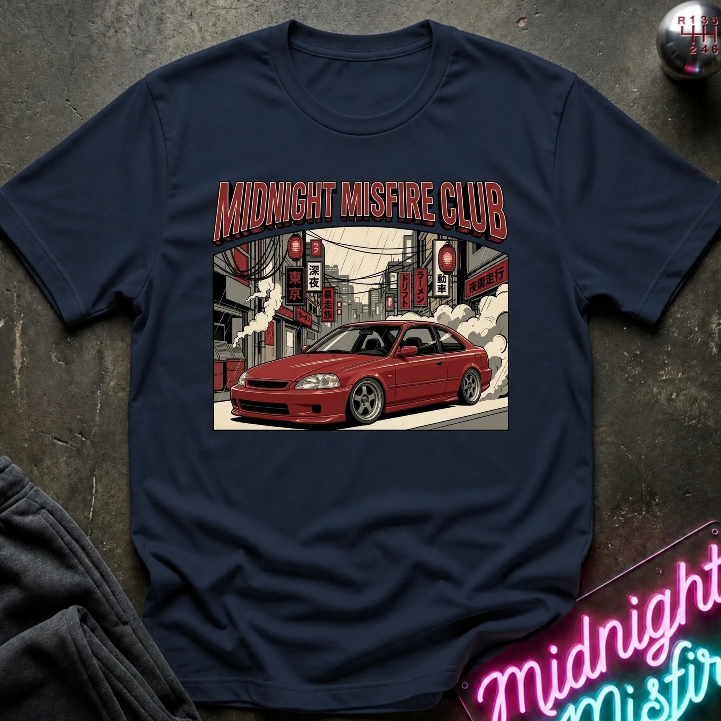 A navy blue graphic t-shirt laid flat on a dark textured surface. The shirt features a central graphic of a red coupe car in a street scene with the text 'MIDNIGHT MISFIRE CLUB' in red.
