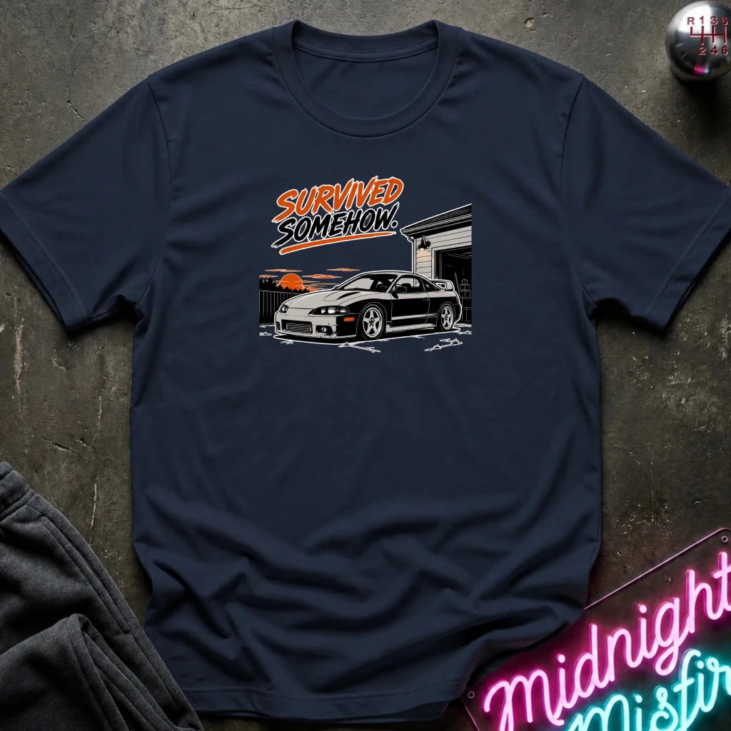 A navy blue graphic t-shirt laid flat on a dark textured surface. The shirt features a graphic of a white sports car in front of a garage with orange and white text reading 'SURVIVED SOMEHOW'.