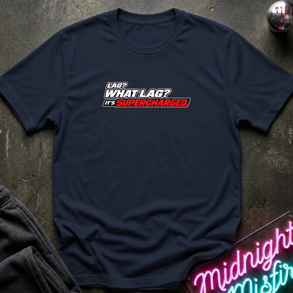 A navy blue short-sleeve t-shirt with a graphic text print reading 'LAG? WHAT LAG? IT'S SUPERCHARGED' in white, black, and red, laid flat on a dark textured surface with a gray sweatshirt and a neon sign partially visible as props.