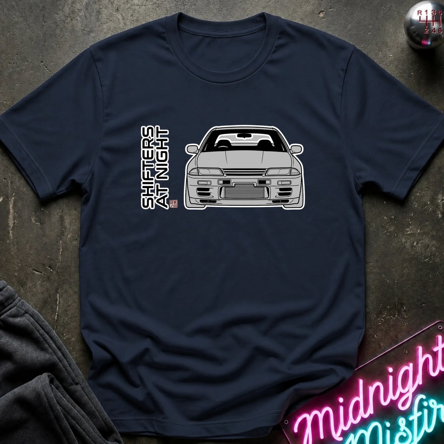 A navy blue short-sleeve t-shirt laid flat on a dark textured surface, featuring a black line art graphic of a sports car and the vertical text 'SHIFTERS AT NIGHT' with a small red and blue flag detail. A neon sign and dark clothing are partially visible as props.