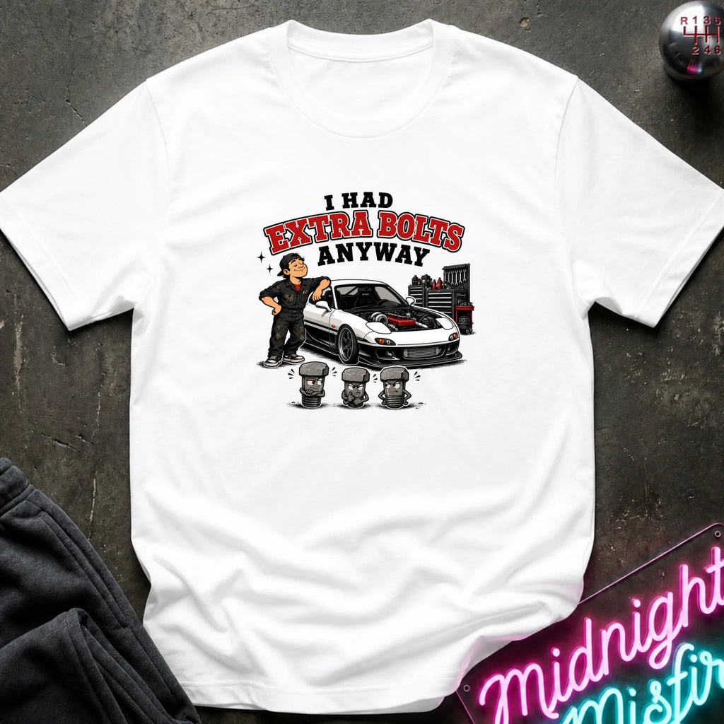 A white short-sleeve graphic t-shirt laid flat on a dark textured surface. The graphic features a cartoon of a man standing next to a white sports car with red detailing, with the text 'I HAD EXTRA BOLTS ANYWAY' in red and black.