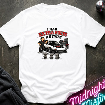 A white short-sleeve graphic t-shirt laid flat on a dark textured surface. The graphic features a cartoon of a man standing next to a white sports car with red detailing, with the text 'I HAD EXTRA BOLTS ANYWAY' in red and black.
