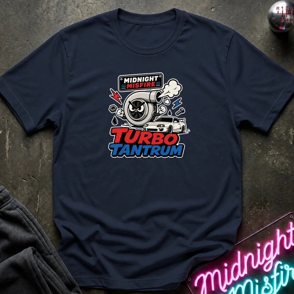 A navy blue graphic t-shirt laid flat on a dark textured surface. The shirt features a central graphic of a turbocharger, a white car, and the text 'MIDNIGHT MISFIRE' and 'TURBO TANTRUM' in red and blue.
