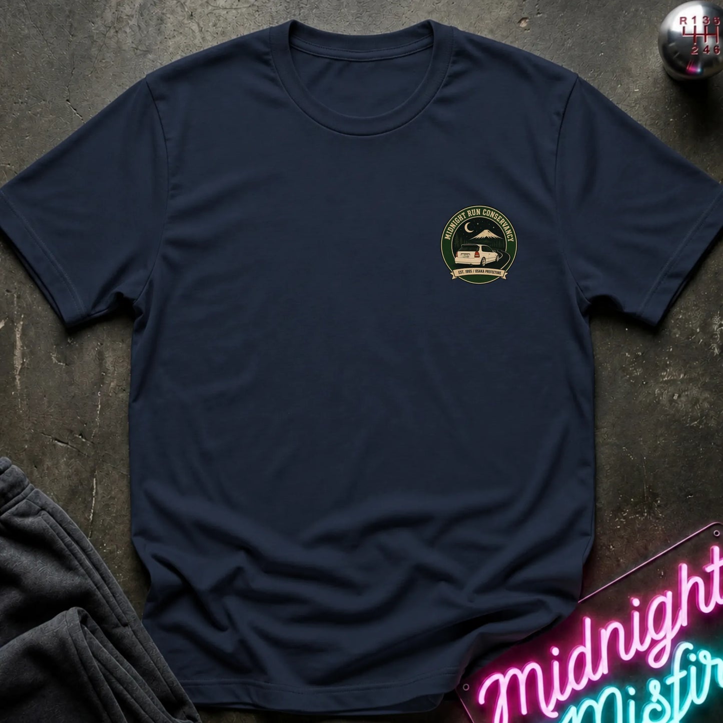 A navy blue short-sleeve crewneck t-shirt with a circular green and black chest graphic, styled flat-lay on a dark textured surface with a neon sign and dark pants nearby.