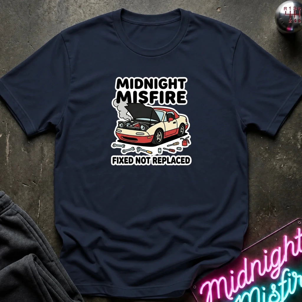 A navy blue graphic t-shirt laid flat on a dark textured surface. The shirt features a graphic of a white and red sports car with its hood open, surrounded by tools, with the text 'MIDNIGHT MISFIRE' above and 'FIXED NOT REPLACED' below.