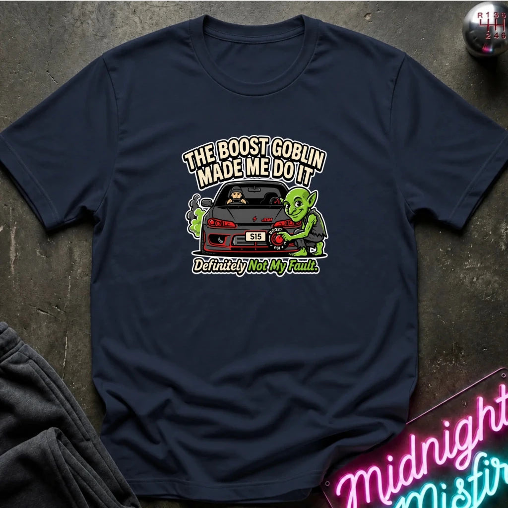 A navy blue short-sleeve graphic t-shirt laid flat on a dark textured surface. The graphic features a green cartoon character next to a red sports car with the text 'THE BOOST GOBLIN MADE ME DO IT' above and 'Definitely Not My Fault.