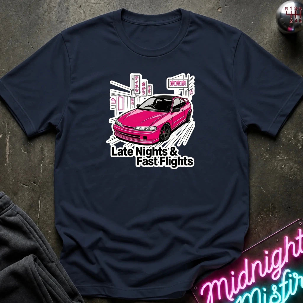 A navy blue graphic t-shirt laid flat on a dark textured surface. The shirt features a graphic of a pink car in front of a city skyline with the text 'Late Nights & Fast Flights'.