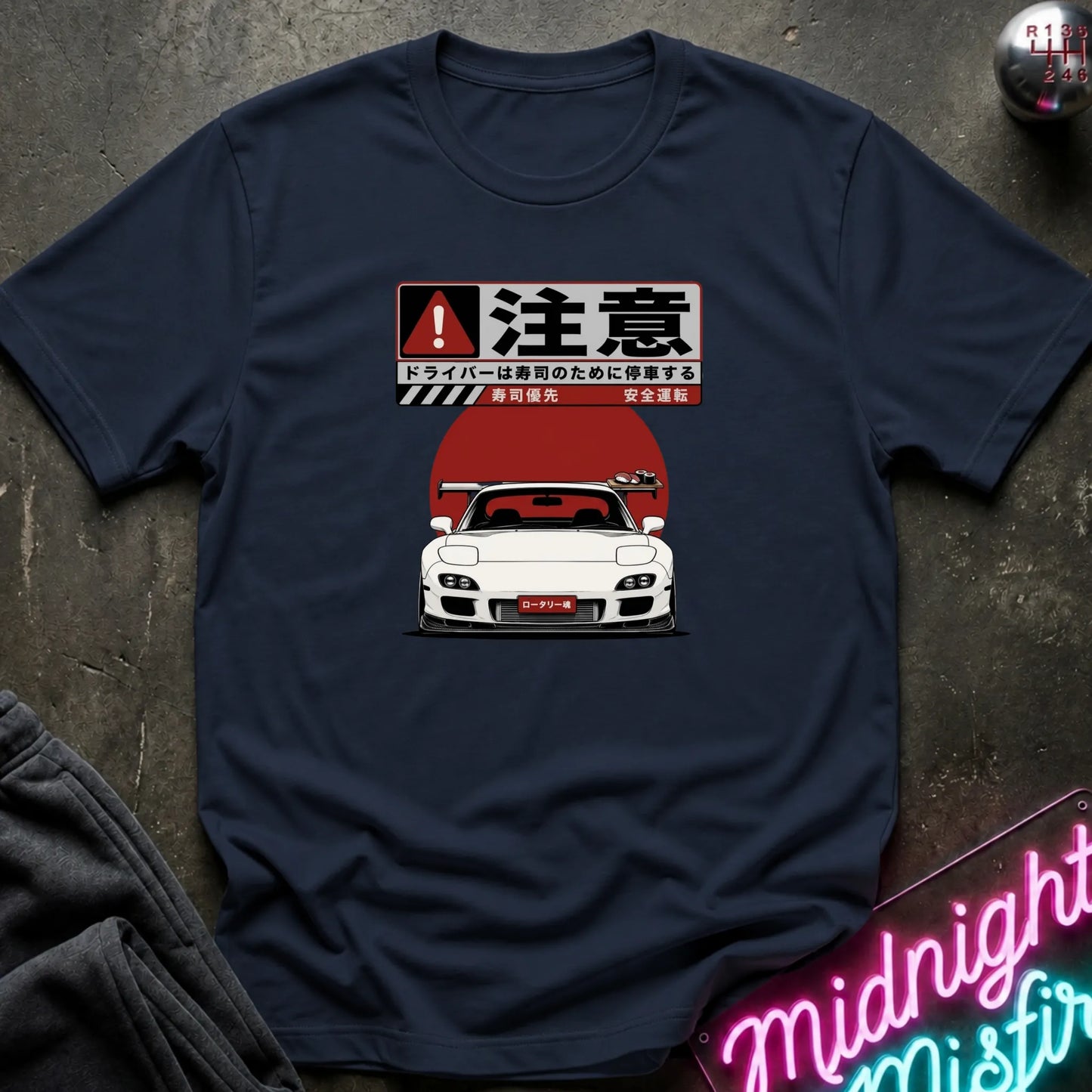 A navy blue short-sleeve t-shirt laid flat on a dark textured surface, featuring a graphic print of a white sports car in front of a red sun with Japanese text and a warning-style sign above it.