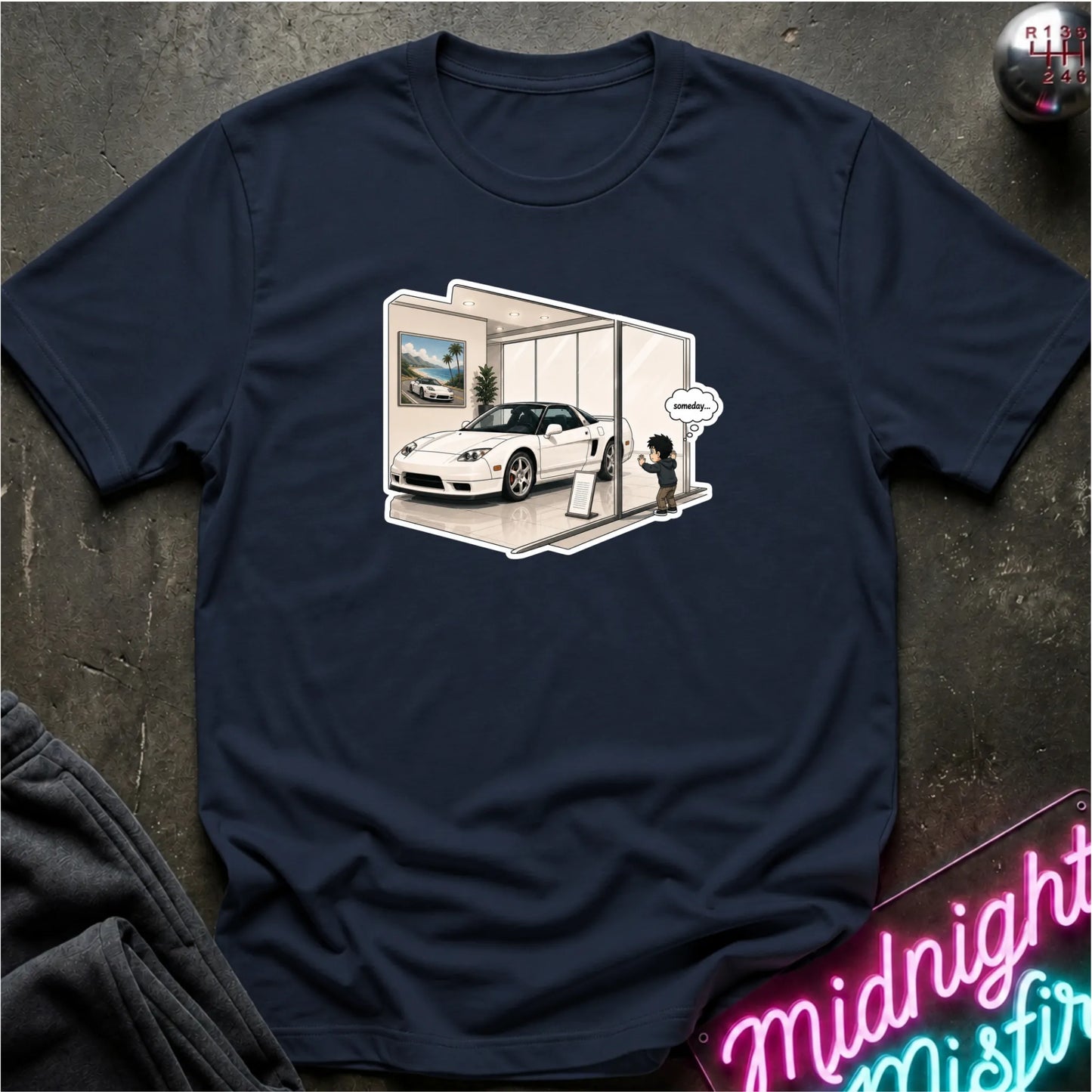 A navy blue short-sleeve t-shirt laid flat on a dark textured surface, featuring a central graphic of a white sports car inside a showroom with a small illustrated person and speech bubble. A neon sign and folded dark fabric are partially visible at the edges.