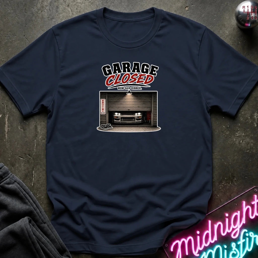 A navy blue graphic t-shirt laid flat on a dark textured surface. The graphic features the text 'GARAGE CLOSED' with a garage door illustration and a car inside.