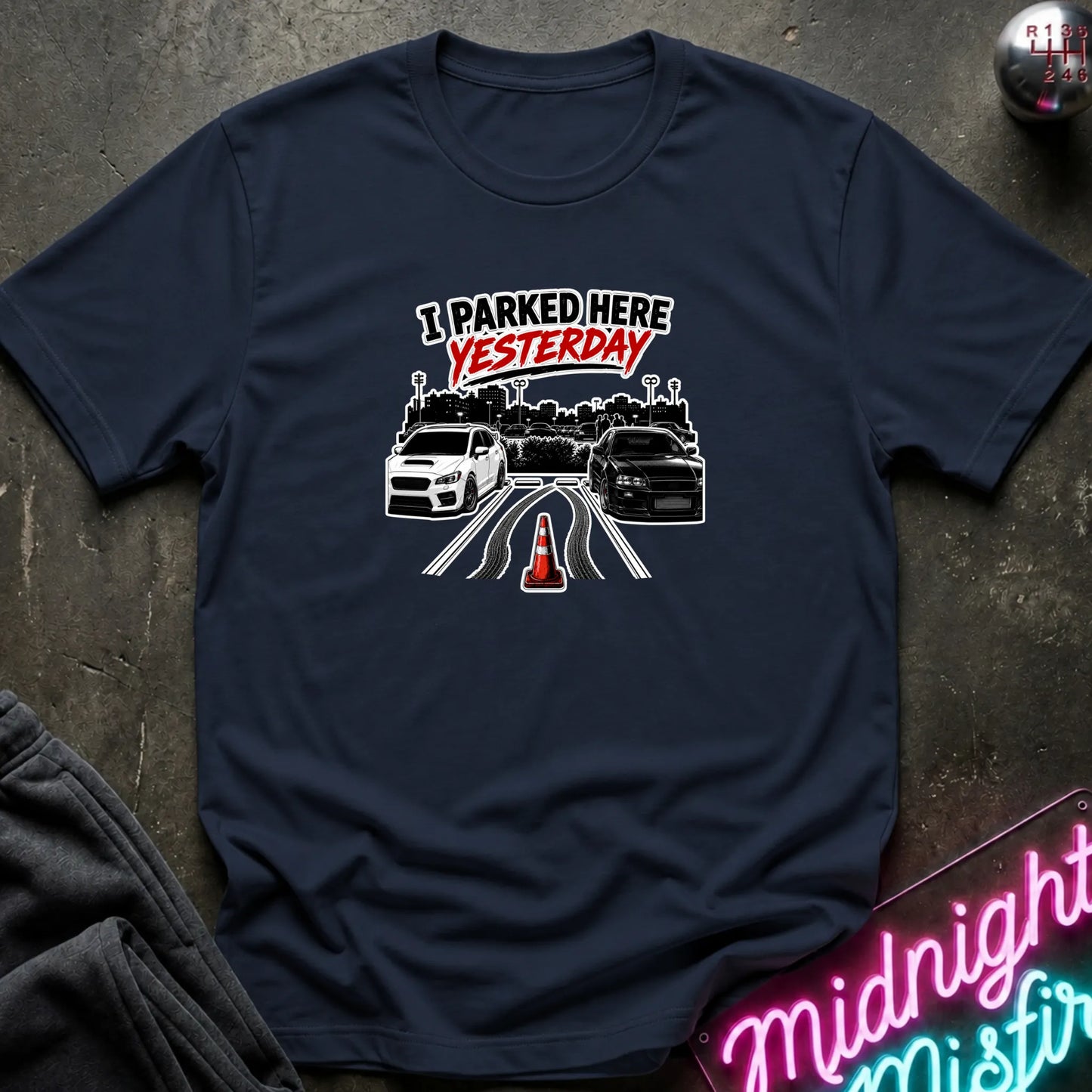 A navy short-sleeved t-shirt  that has 2 cars parked in a space in between empty space with skid marks.
