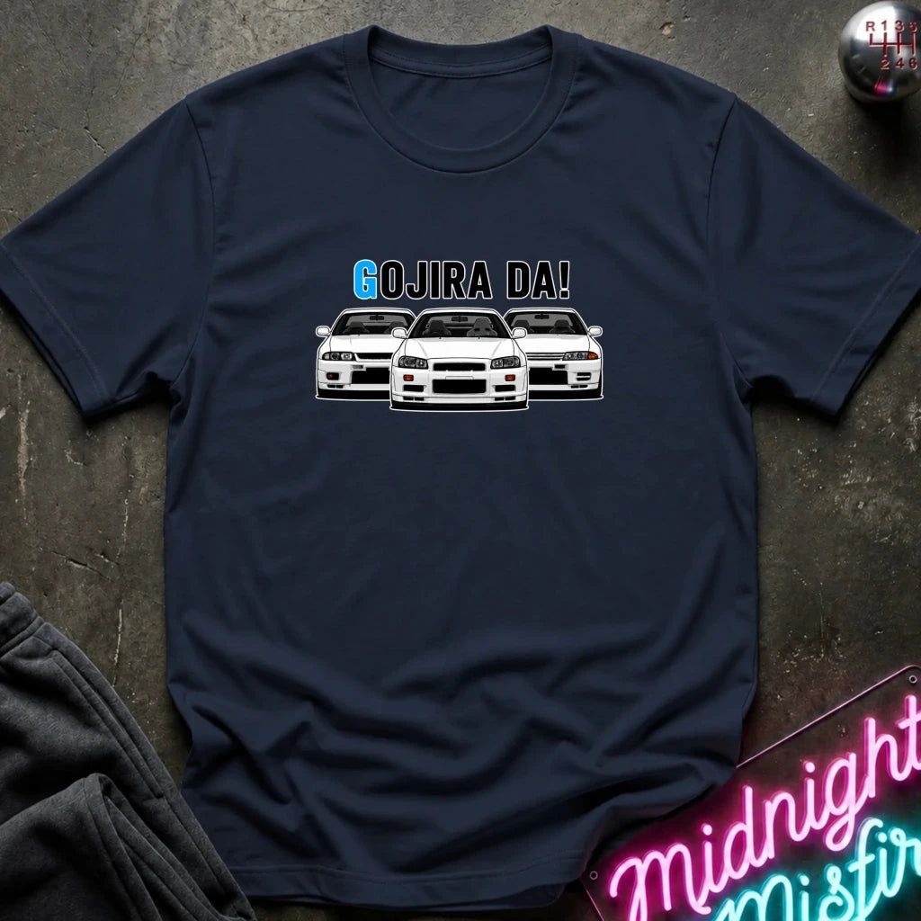 A navy blue short-sleeve t-shirt laid flat on a dark textured surface, featuring a graphic of three white sports cars with the text 'GOJIRA DA!' above them. A neon sign and folded dark fabric are partially visible as props.