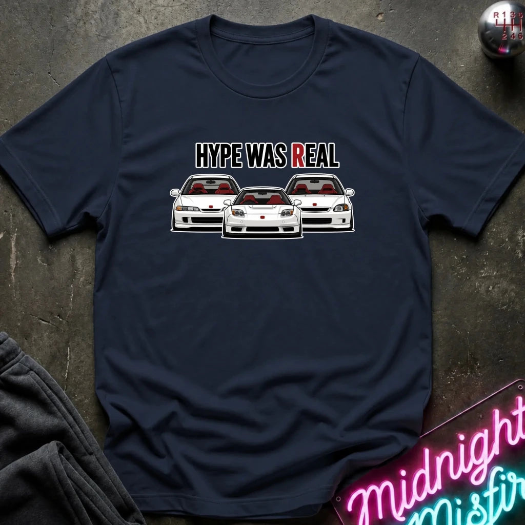 A navy blue short-sleeve t-shirt laid flat on a dark textured surface. The shirt features a graphic print of three white sports cars with red interiors and the text 'HYPE WAS REAL' above them.