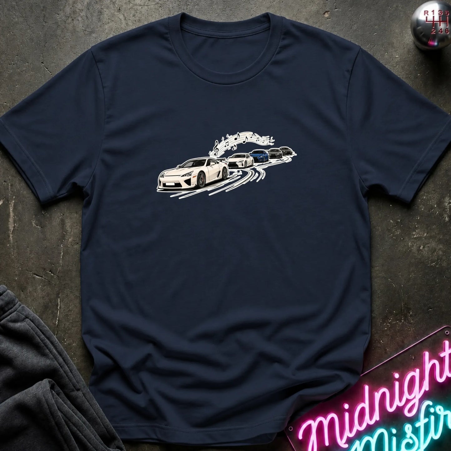 A navy blue short-sleeve crewneck t-shirt featuring a graphic print of a white sports car leading a line of other cars, laid flat on a dark textured surface with a neon sign and dark pants partially visible.