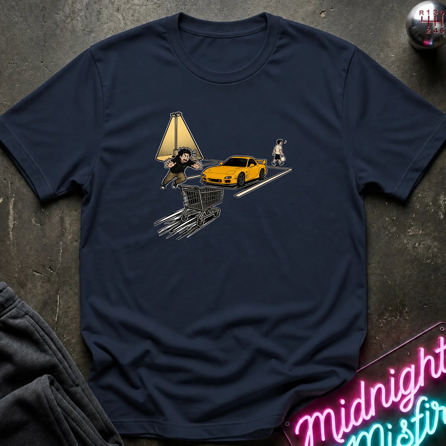 A navy blue short-sleeve t-shirt laid flat on a dark textured surface, featuring a graphic print of a yellow sports car, a shopping cart, a lamp, and a person.