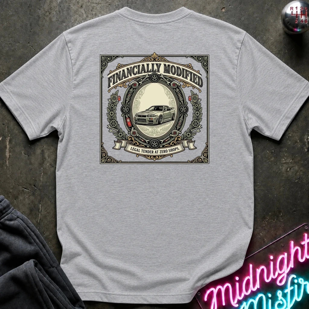 A light gray heather crewneck t-shirt laid flat on a dark textured surface, featuring a large vintage-style graphic on the back with the text 'FINANCALLY MODIFIED' and a car illustration. A neon sign and sweatpants are partially visible as props.