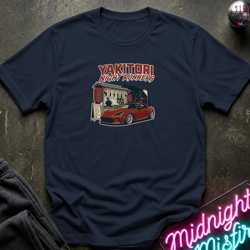 A navy blue graphic t-shirt laid flat on a dark textured surface. The shirt features a red and white graphic print of a sports car in front of a building with the text 'YAKITORI NIGHT RUNERS'.