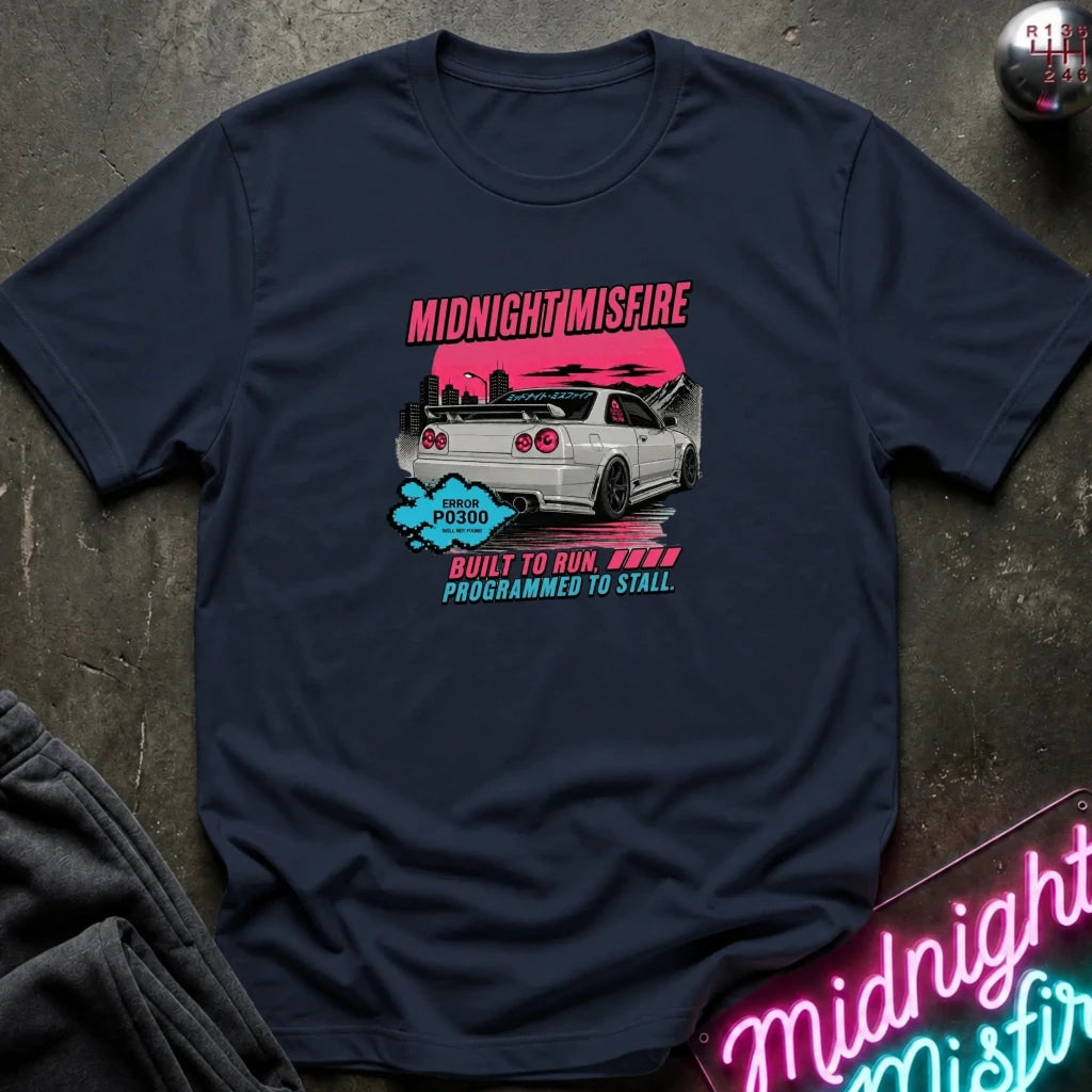 A navy blue graphic t-shirt laid flat on a dark textured surface. The shirt features a pink and blue graphic of a white sports car with the text 'MIDNIGHT MISFIRE' and 'BUILT TO RUN PROGRAMMED TO STALL'.