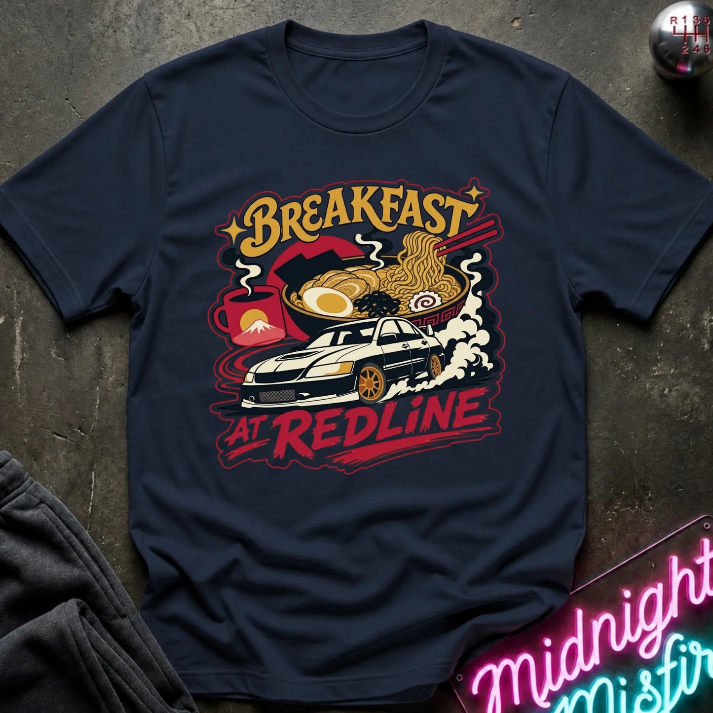 A navy blue graphic t-shirt laid flat on a dark textured surface. The shirt features a colorful retro-style print with the words 'BREAKFAST AT REDLINE' and an illustration of a car, ramen noodles, and an egg.