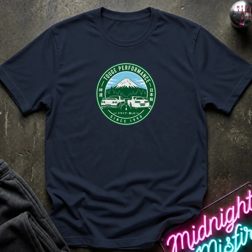 A navy blue short-sleeve t-shirt laid flat on a dark textured surface, featuring a circular green and white graphic on the chest with a mountain, two cars, and the text
