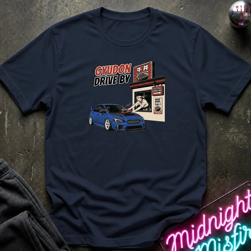 A navy blue short-sleeve graphic t-shirt laid flat on a dark textured surface. The graphic features a blue sports car in front of a Japanese-style shop with the text 'GYUDON DRIVE BY'.