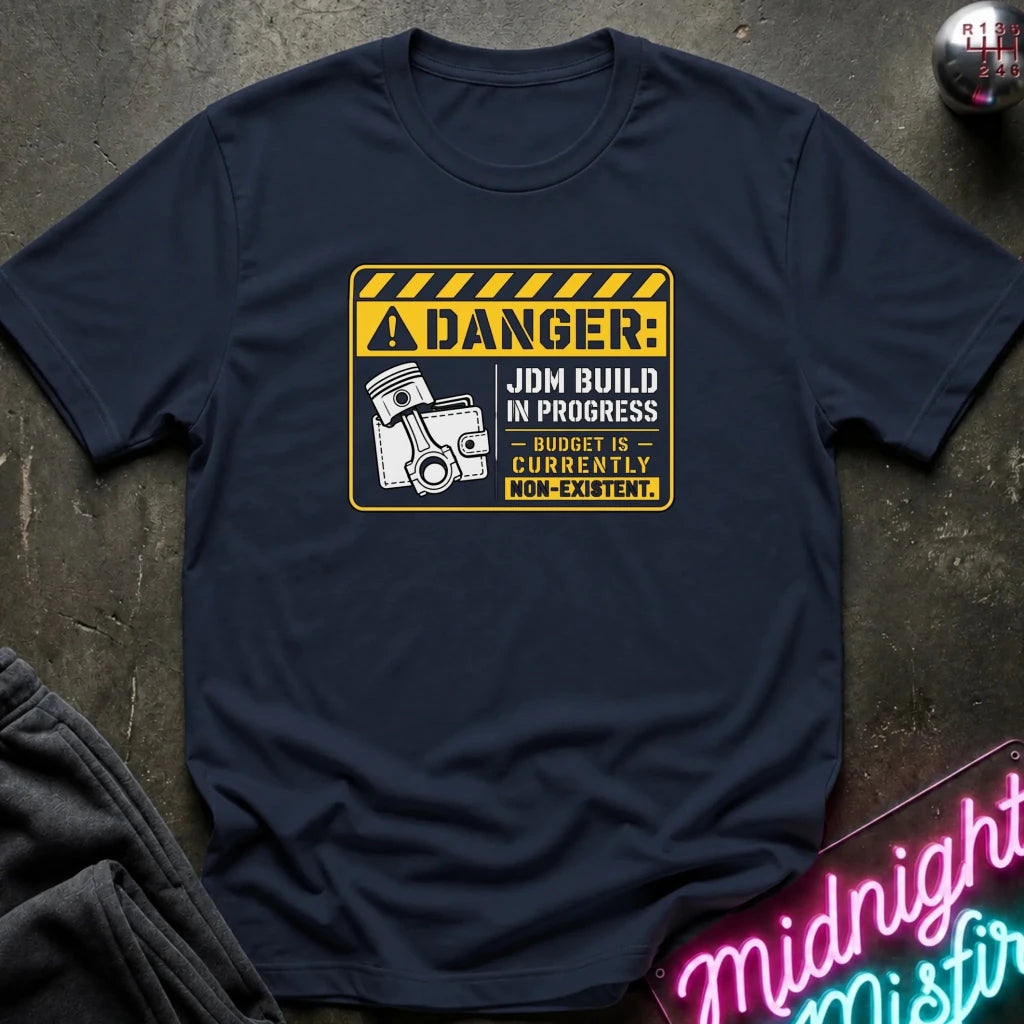 A navy blue crewneck t-shirt with a yellow and black warning sign graphic on the chest that reads 'DANGER: JDM BUILD IN PROGRESS - BUDGET IS CURRENTLY NON-EXISTENT.' The shirt is styled flat on a dark textured surface with a neon sign and folded dark fabric in the corners.