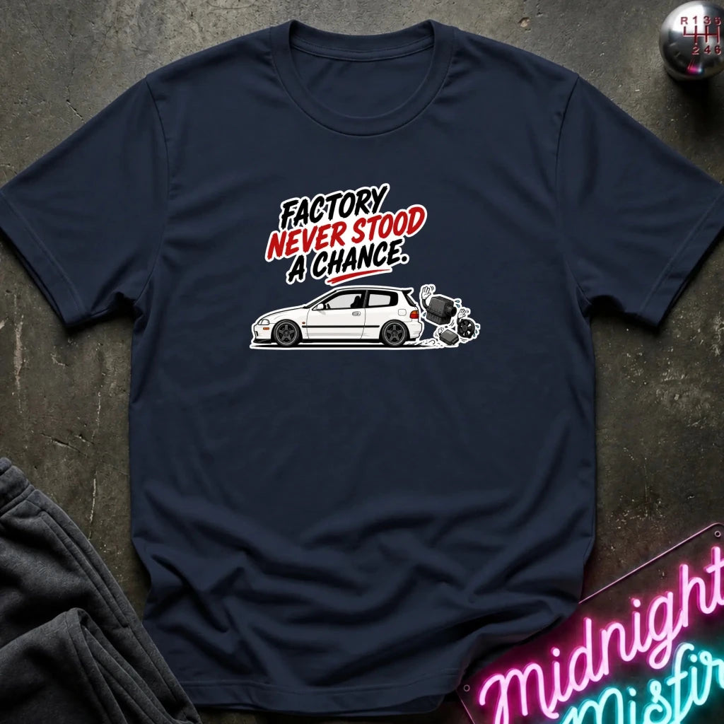 A navy blue graphic t-shirt laid flat on a dark textured surface. The shirt features a white car illustration and the text 'FACTORY NEVER STOOD A CHANCE' in white and red.