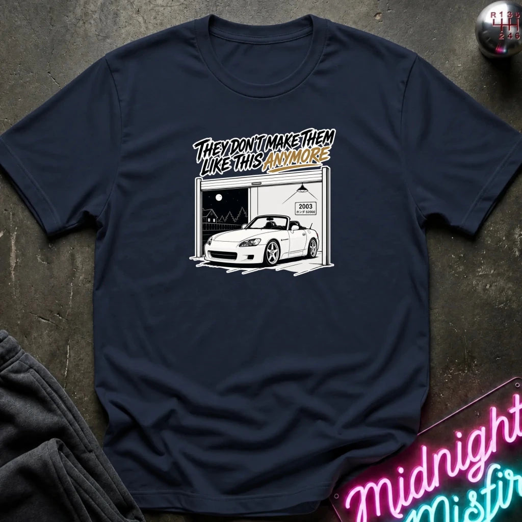 A navy blue short-sleeve t-shirt with a graphic print of a white sports car in a garage and the text 'THEY DON'T MAKE THEM LIKE THIS ANYMORE' in a stylized font. The shirt is laid flat on a dark textured surface with a neon sign and dark clothing partially visible in the corners.