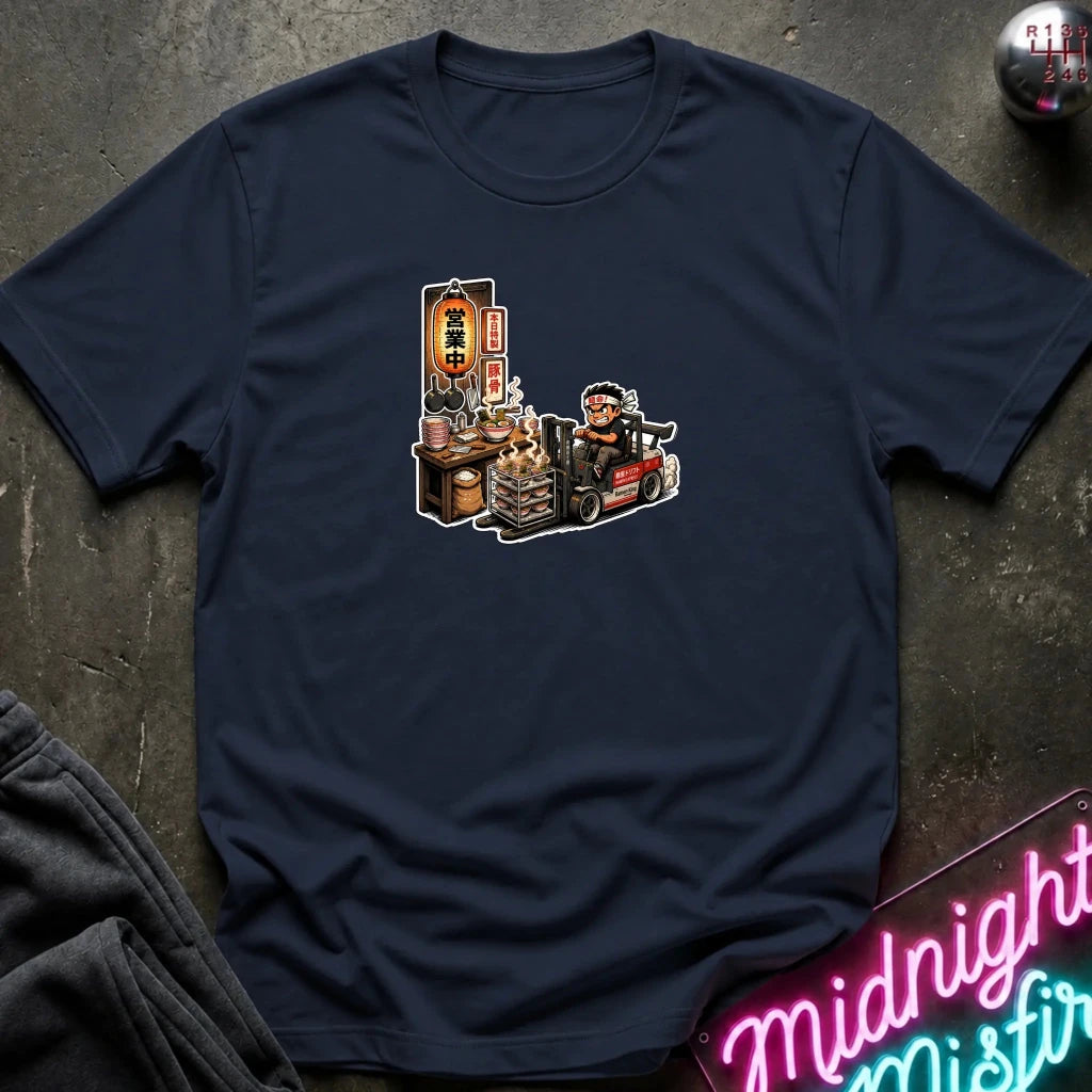 A navy blue short-sleeve t-shirt with a cartoon graphic of a street food vendor on a forklift, laid flat on a dark textured surface with a neon sign and folded sweatpants nearby.