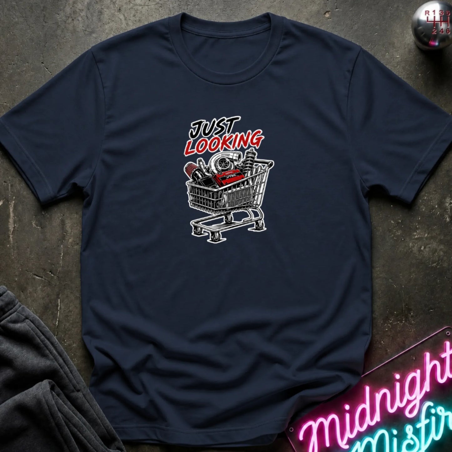 A navy short-sleeve t-shirt laid flat on a dark textured surface, featuring a graphic print of a shopping cart filled with items and the text 'JUST LOOKING' in black and red. Partially visible props include dark pants in the lower left and a neon-style sign in the lower right.