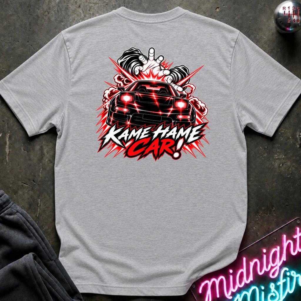 A light gray heather t-shirt laid flat on a dark textured surface, featuring a large graphic print of a black sports car with red headlights and the text 'KAME HAME CAR!' in white and red.