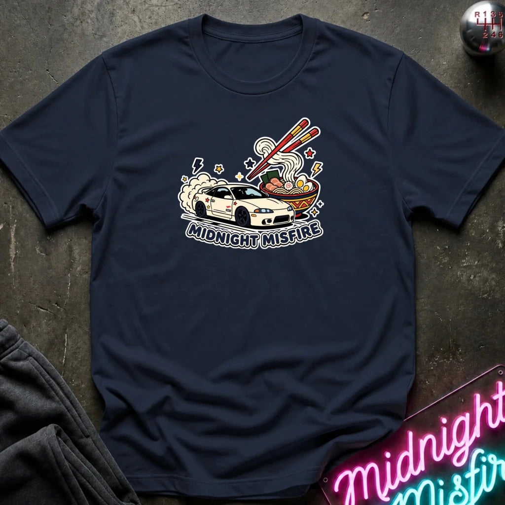 A navy blue short-sleeve t-shirt laid flat on a dark textured surface, featuring a graphic of a white sports car driving into a ramen bowl with chopsticks, and the text 'MIDNIGHT MISFARE' printed below. A neon sign with the same text is partially visible in the bottom right corner.