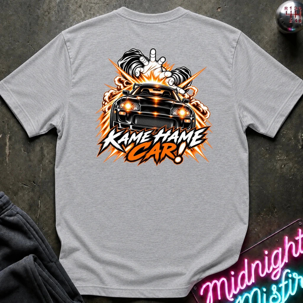 A light gray heather t-shirt laid flat on a dark textured surface, featuring a large graphic print on the back with a black car, flames, and the text 'KAME HAME CAR!'.