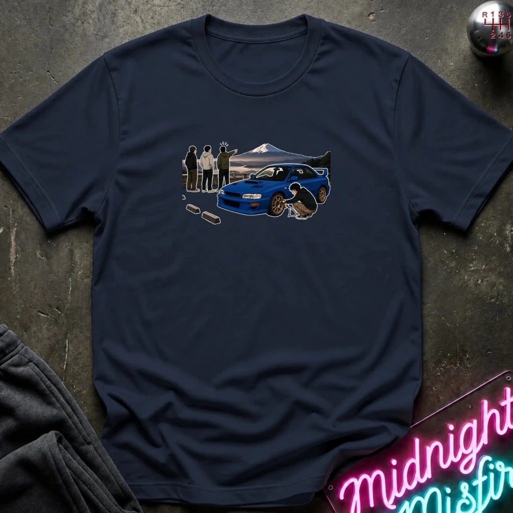 A navy blue short-sleeve t-shirt laid flat on a dark textured surface, featuring a graphic print of a blue sports car, a mountain, and a group of people. 