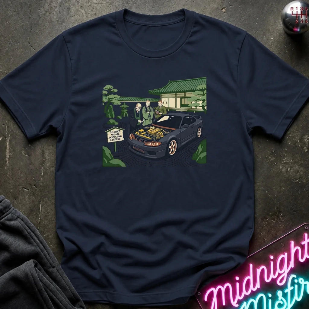 A navy blue short-sleeve t-shirt laid flat on a dark textured surface, featuring a colorful graphic print of a black sports car with its hood open, surrounded by greenery and a house. A folded dark garment and a neon sign are partially visible at the edges.
