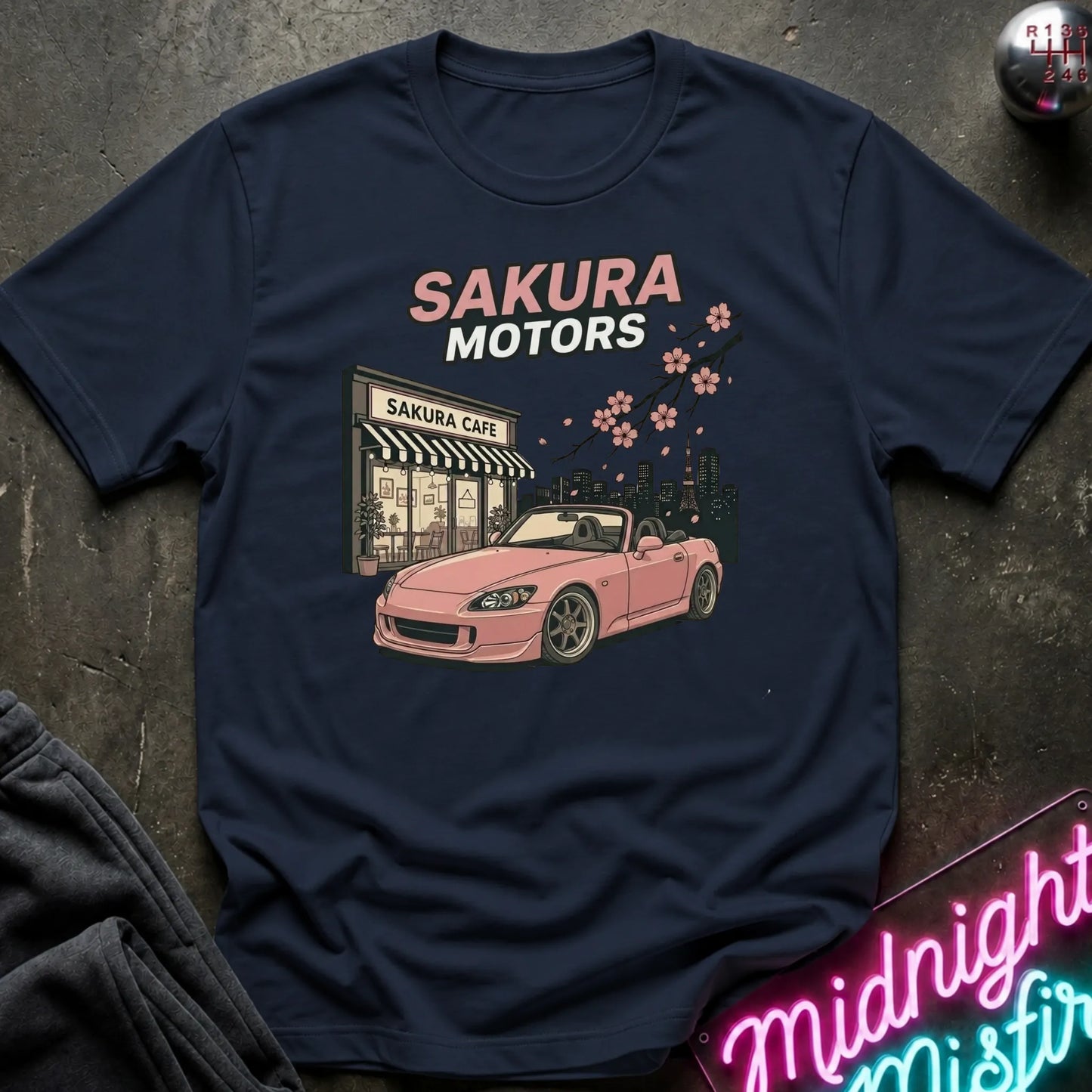 navy short-sleeve graphic T-shirt laid flat on a dark textured surface. The shirt features a printed design of a pink convertible sports car in front of a building labeled 'Sakura Cafe', with 'SAKURA MOTORS' text above and cherry blossoms across the top.
