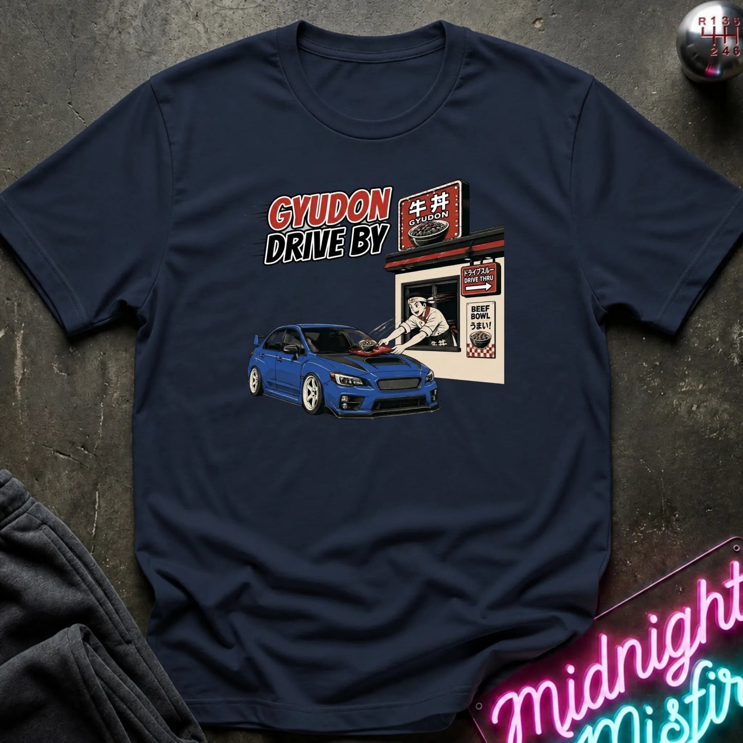 A navy blue graphic t-shirt laid flat on a dark textured surface. The graphic features a blue sports car and a Japanese restaurant sign with the text 'GYUDON DRIVE BY'.