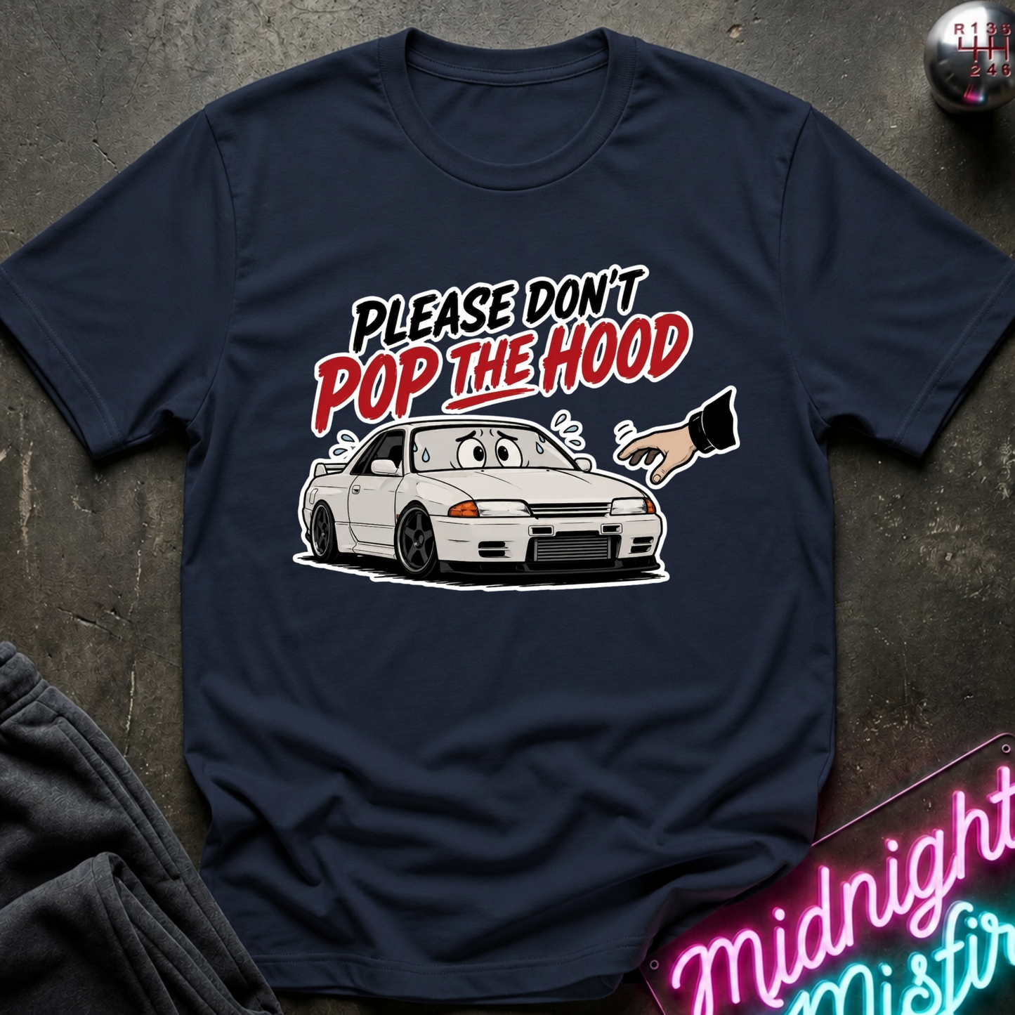 A navy blue graphic t-shirt laid flat on a dark textured surface. The shirt features a cartoon illustration of a white sports car with a surprised face, a hand pointing toward it, and the text 'PLEASE DON'T POP THE HOOD' in white and red lettering.