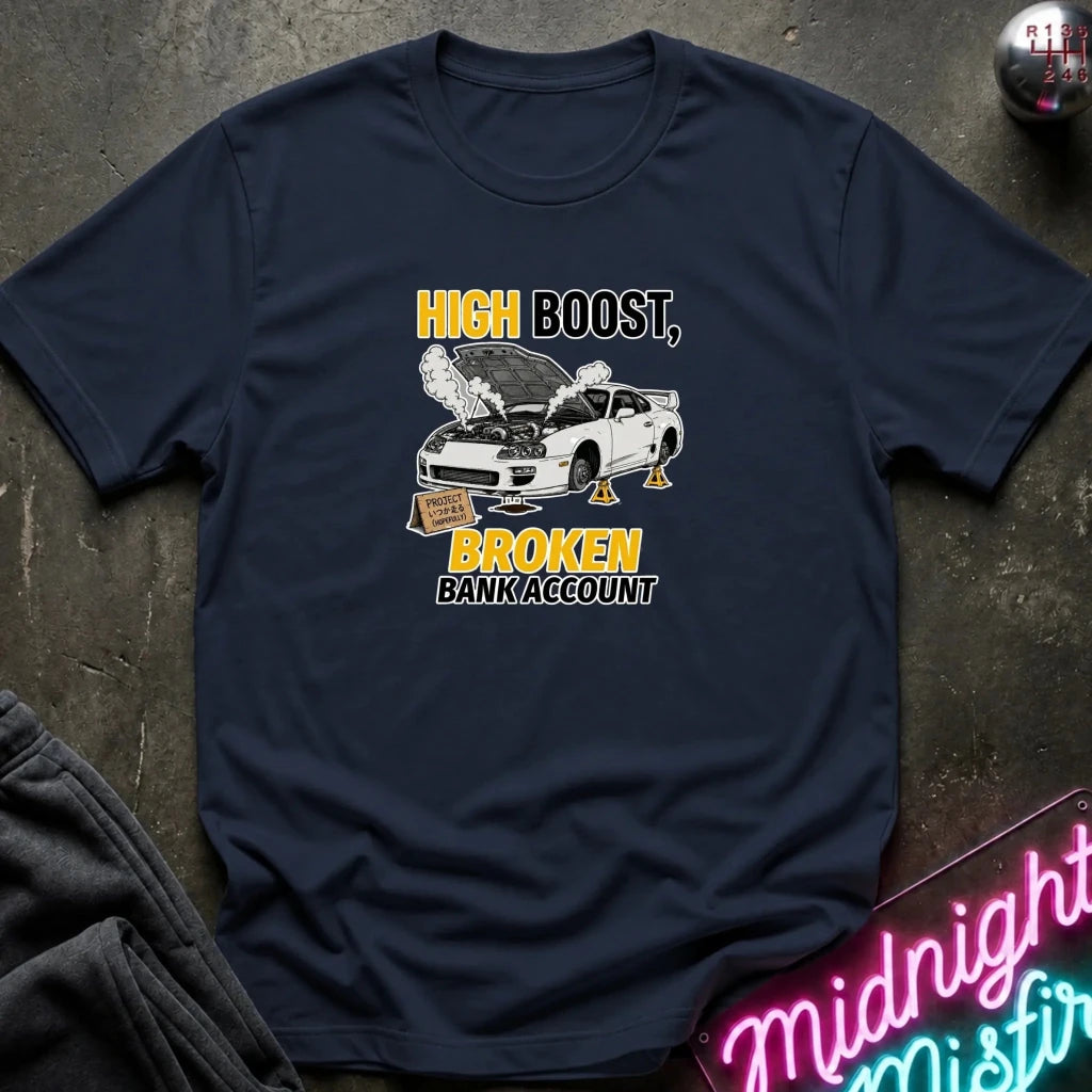 A navy blue graphic t-shirt laid flat on a dark textured surface. The shirt features a white car with its hood open, emitting smoke, with the text 'HIGH BOOST, BROKEN BANK ACCOUNT' in yellow and white.