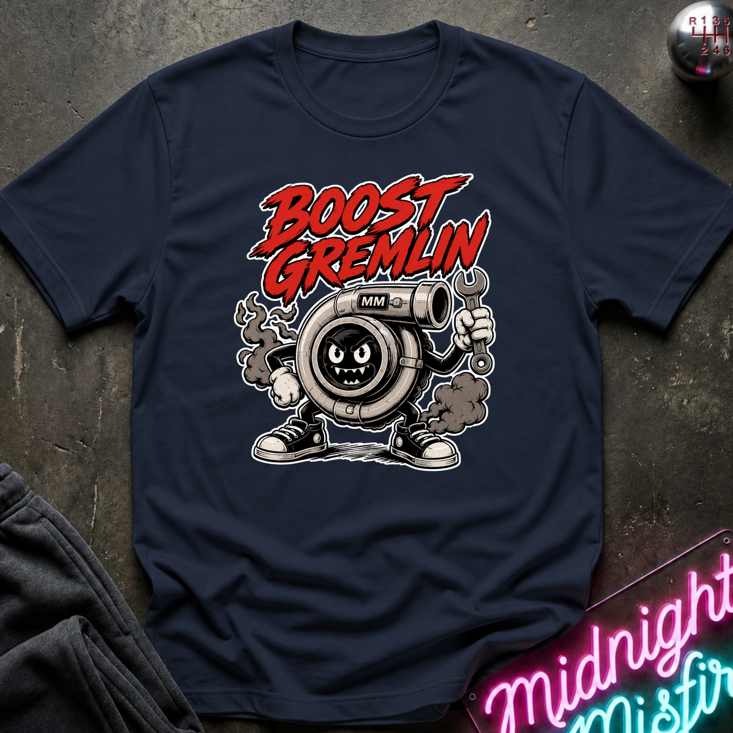 A navy blue graphic t-shirt featuring a cartoon turbocharger character with the text 'BOOST GREMLIN' in red and white. The shirt is laid flat on a dark textured surface with a neon sign and dark sweatpants partially visible.