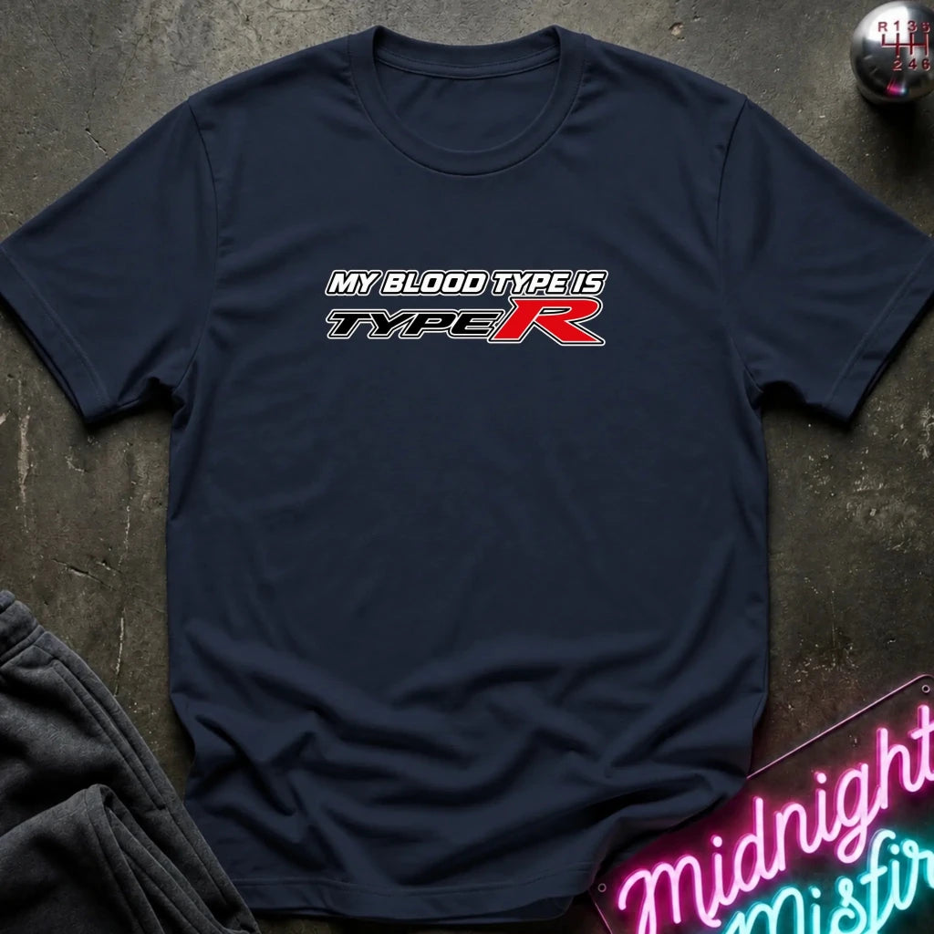 A navy blue short-sleeve t-shirt with a 'MY BLOOD TYPE IS TYPE R' graphic printed on the chest, laid flat on a dark textured surface with a neon sign and clothing props.