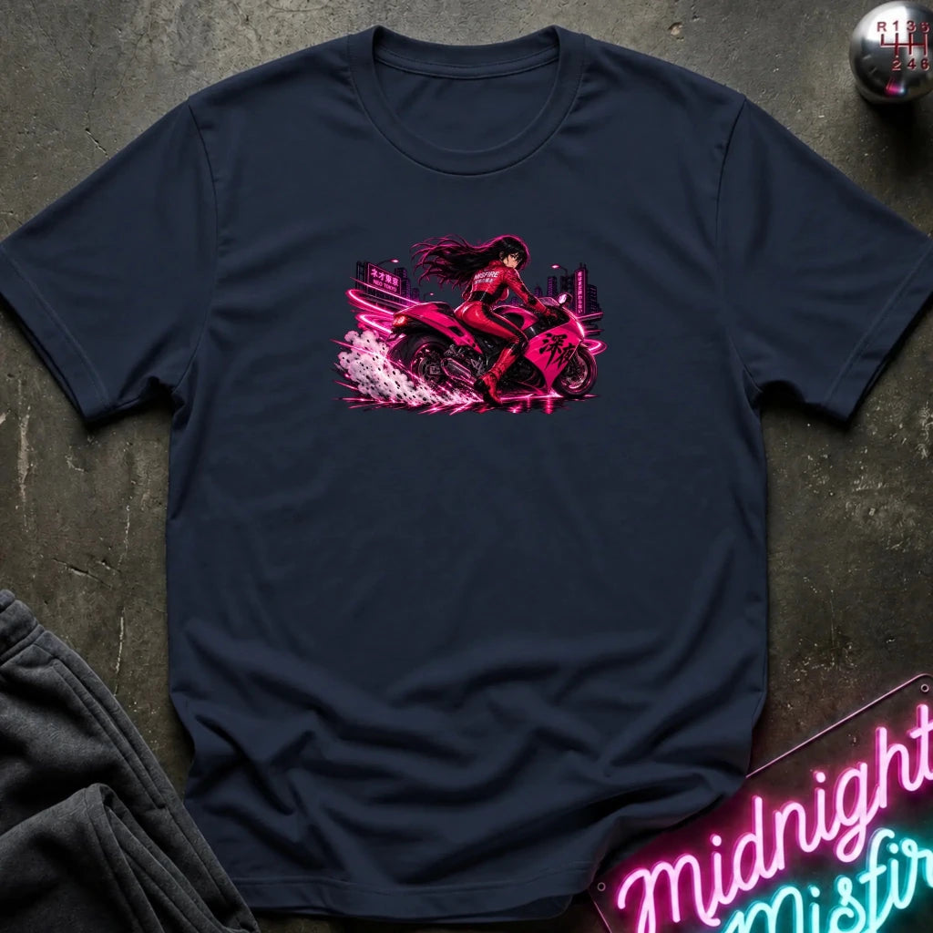 A navy blue short-sleeve t-shirt laid flat on a dark textured surface, featuring a vibrant pink and red graphic of a woman riding a sport motorcycle. The shirt is styled with a folded pair of dark sweatpants, a metallic sphere, and a neon sign in the background.